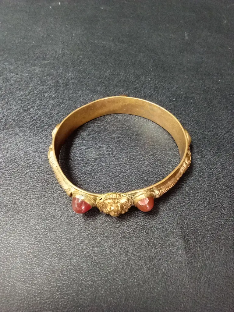 ANTIQUE-STYLE GILT METAL BANGLE WITH CENTRAL LION MASK, FLANKED BY RED GLASS CABOCHONS AND ROPE-TWIST DETAIL Image 1++