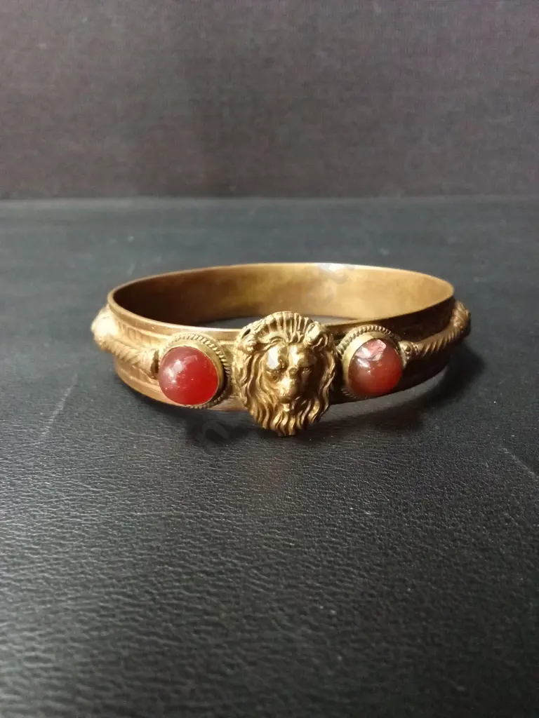 ANTIQUE-STYLE GILT METAL BANGLE WITH CENTRAL LION MASK, FLANKED BY RED GLASS CABOCHONS AND ROPE-TWIST DETAIL Image 1++