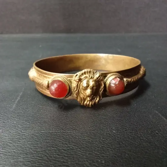 ANTIQUE-STYLE GILT METAL BANGLE WITH CENTRAL LION MASK, FLANKED BY RED GLASS CABOCHONS AND ROPE-TWIST DETAIL
